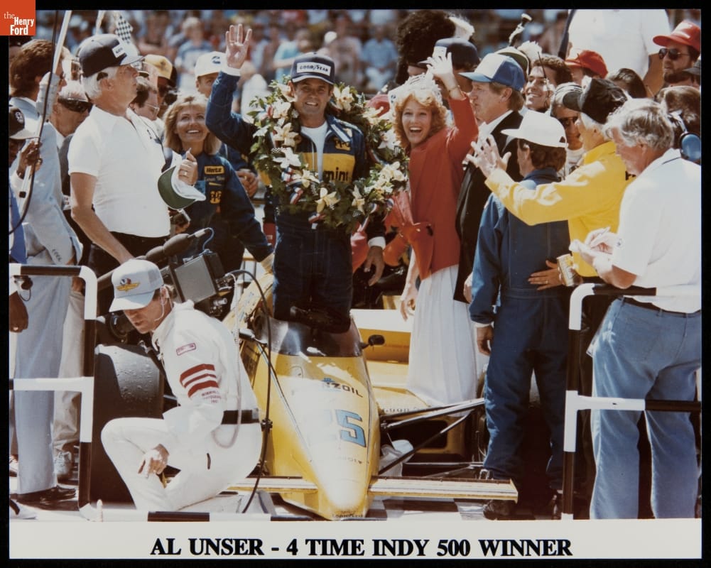 Al Unser, Sr., four-time Indy 500 winner Man in jumpsuit with wreath around neck stands in a race car waving to the camera with a crowd of people looking on