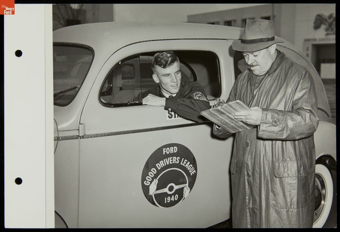 Man in raincoat and hat holds a paper by a car with decal with text on side; young man sticks head and elbow out driver side window
