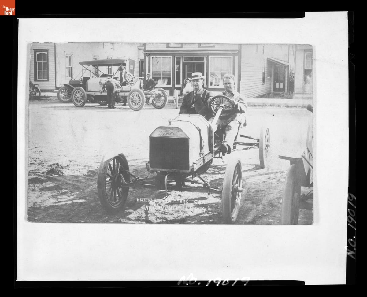Frank Kulick Driving the Ford Special, Winner of the Algonquin Hill Climb, Illinois, 1912 Black-and-white photo of two men sitting in an early open car on a street with other cars, people, and buildings in the background