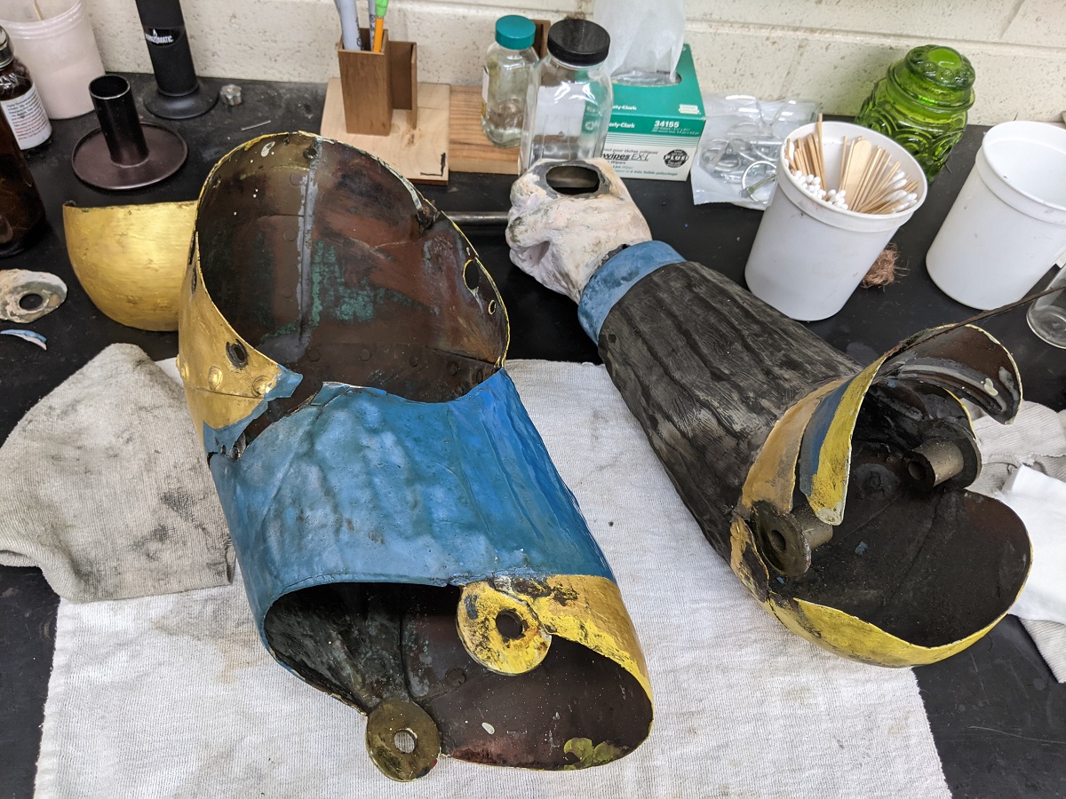 Disassembly of Magog’s arm Painted metal pieces, one in the shape of a forearm and hand, on a cloth on a workbench