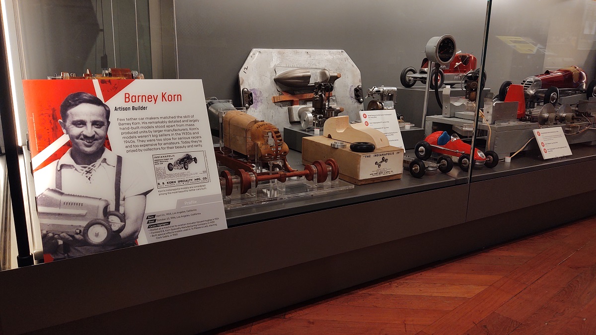 Barney Korn display in Driven to Win in Henry Ford Museum of American Innovation Glass display case containing a sign with text and images and model cars, both whole and in parts and pieces