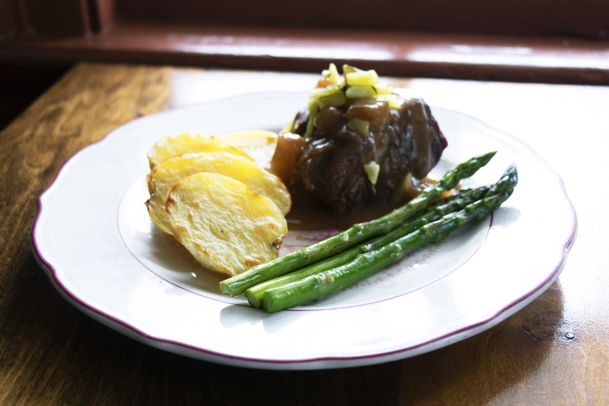Eagle Tavern’s Bouilli Beef White plate filled with beef with gravy topped with chopped vegetables, scalloped potatoes, and asparagus, sitting on wooden table
