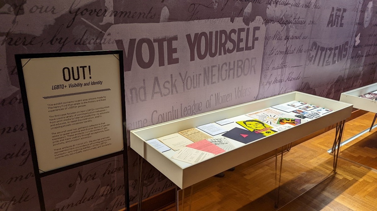 OUT!: LGBTQ+ Visibility and Identity exhibit in Henry Ford Museum of American Innovation A display case holds paper items by a graphics-covered wall and a sign with text