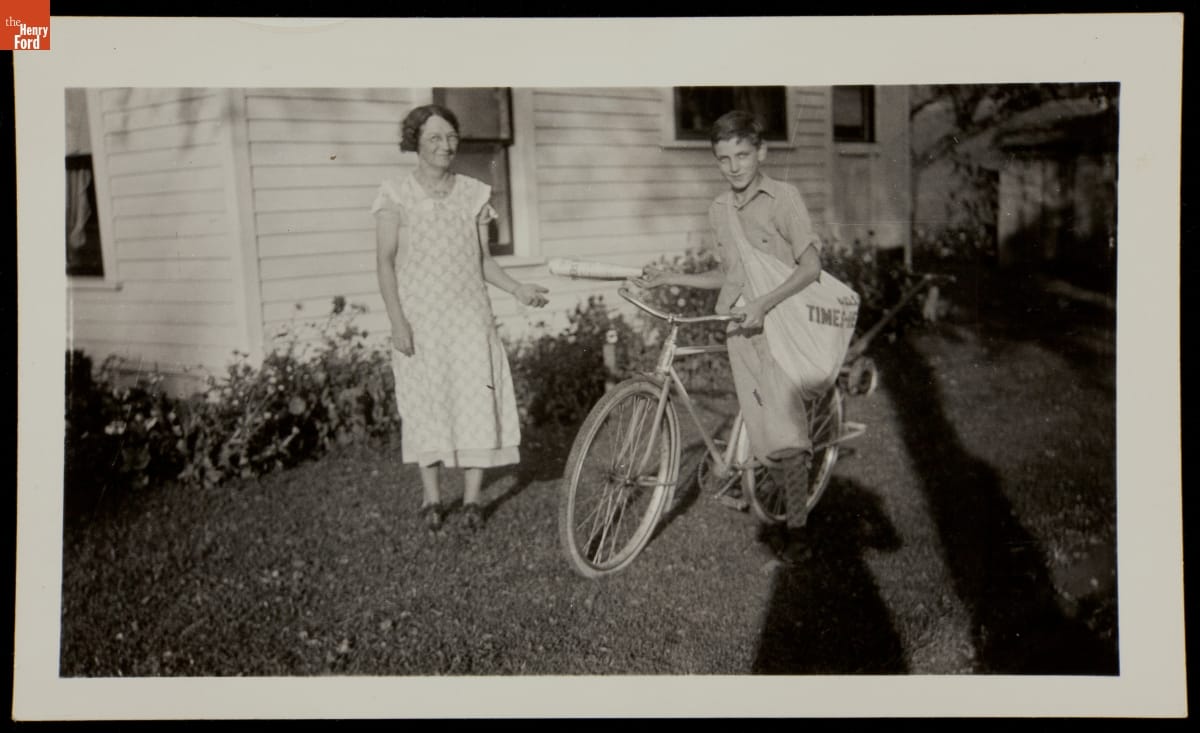 A paperboy standing next to a bicycle with a woman to his right