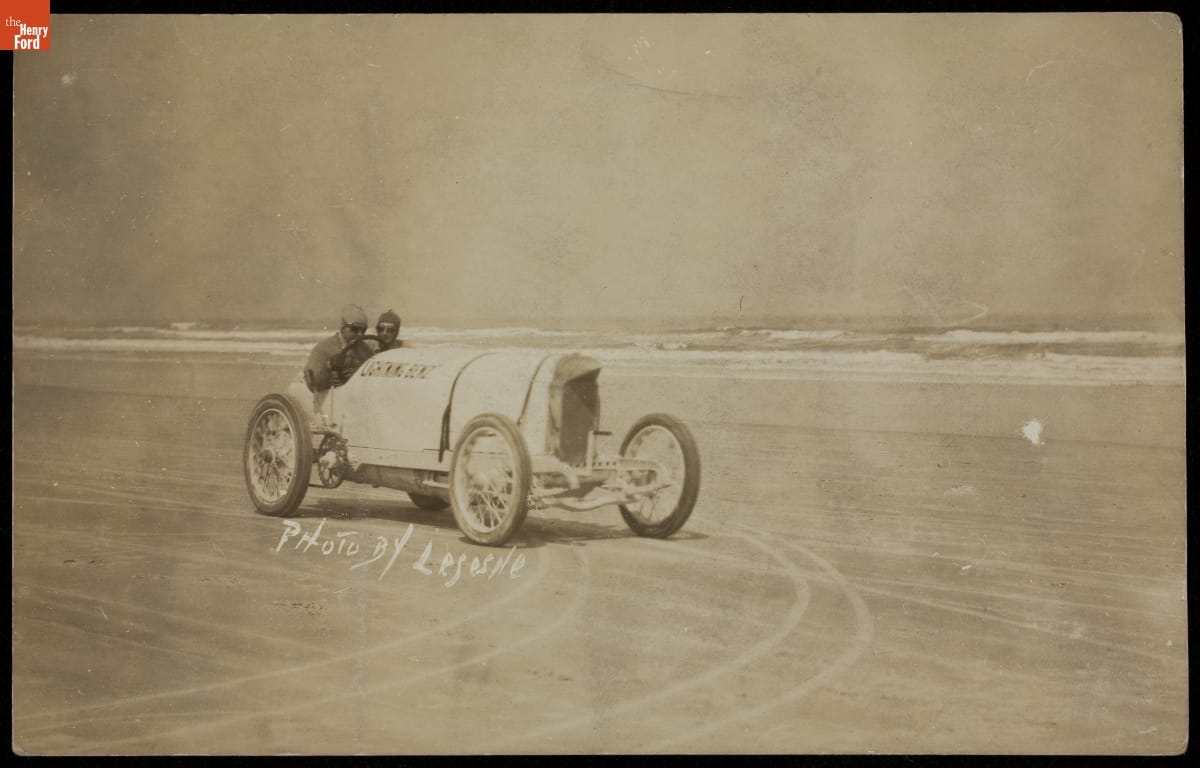 Barney Oldfield in Sepia-tone photo of two men in early open race car on beach with ocean behind them; also contains text