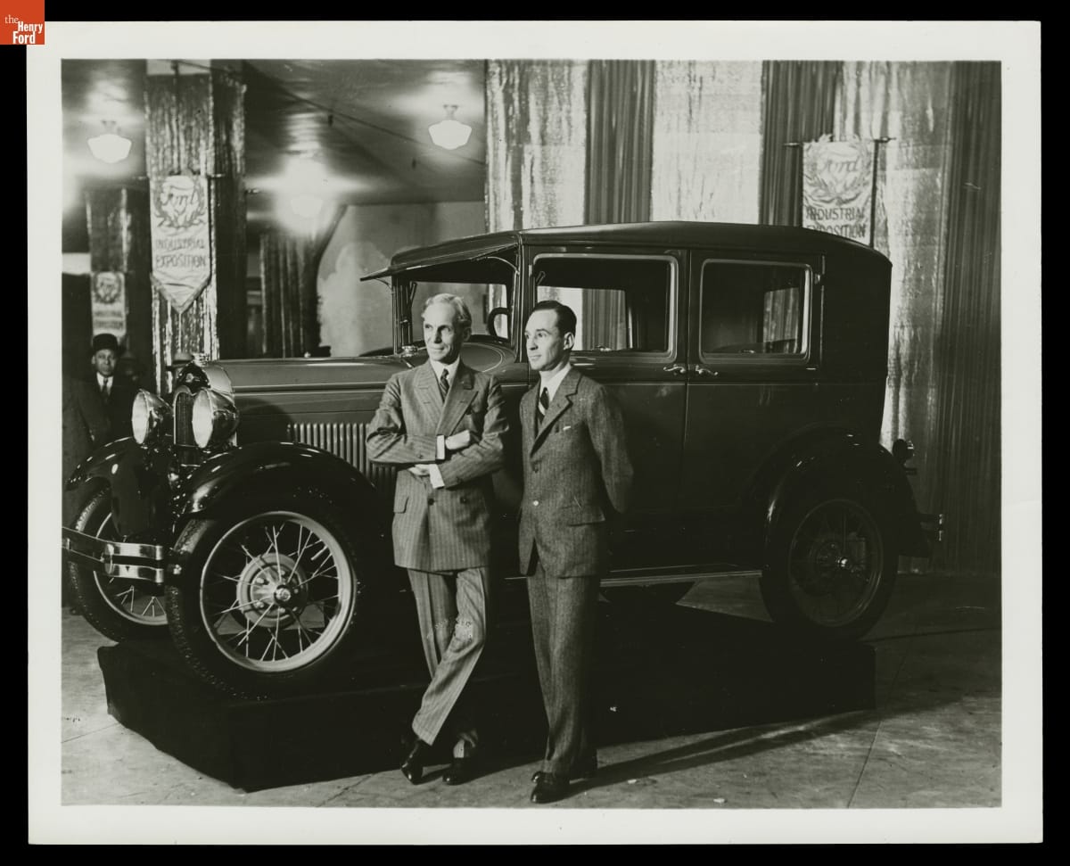 Henry Ford and Edsel Ford with Ford Model A Fordor Sedan, New York Industrial Exposition, 1928 Two men wearing suits stand by car in indoor space with lights and banners